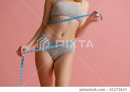 Cropped portrait of beautiful, natural female body in gray inner wear, measuring waist isolated over pink background. Body positivity Cropped portrait of beautiful, natural female body in gray inner wear, measuring waist isolated over pink background. Body positivity 83273624