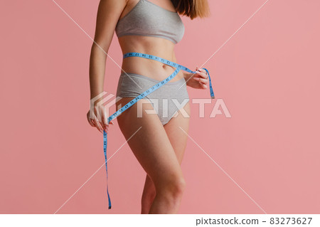 Cropped portrait of beautiful, natural female body in gray inner wear, measuring waist isolated over pink background. Body positivity Cropped portrait of beautiful, natural female body in gray inner wear, measuring waist isolated over pink background. Body positivity 83273627