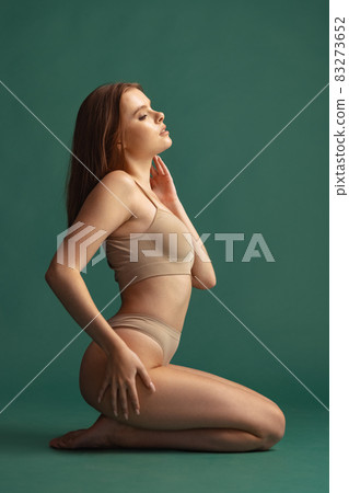 Full-length portrait of beautiful woman sitting in beige cotton underwear isolated over green background. Side view Full-length portrait of beautiful woman sitting in beige cotton underwear isolated over green background. Side view 83273652
