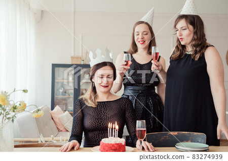 Three women friends celebrating 30s birthday together gathering at table at home with wine and birthday cake with candles. Women in party hat and birthday girl in crown blows out candles. 83274939