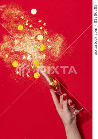 Female hand holding champagne flute with golden confetti isolated over red background 83280388