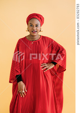 Women in traditional African style. Waist up studio shot of beautiful African woman in red dress, headscarf and jewelry posing against yellow background and looking at camera. 83282783