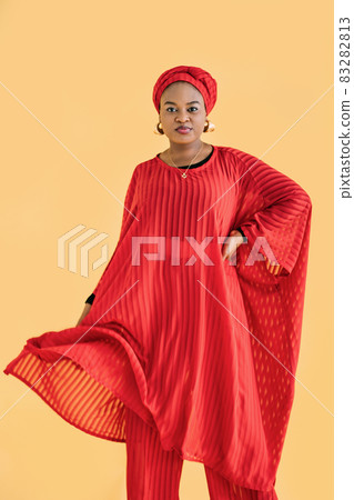 Portrait of charming pleasant African woman wearing red traditional red suit and turban, looking at camera isolated on yellow color background. Ethnic people and traditional fashion. 83282813