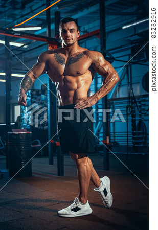 Portrait of young man, muscled athlete, bodybuilder posing at sport gym, indoors. Concept of sport, activity, healthy lifestyle Portrait of young man, muscled athlete, bodybuilder posing at sport gym, indoors. Concept of sport, activity, healthy lifestyle 83282916