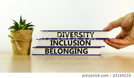 Diversity, inclusion and belonging symbol. Books with words 'diversity, inclusion and belonging' on beautiful white background. Male hand, house plant. Diversity, business, inclusion and belonging Diversity, inclusion and belonging symbol. Books with words 'diversity, inclusion and belonging' on beautiful white background. Male hand, house plant. Diversity, business, inclusion and belonging 83284228