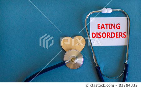 Eating disorders symbol. White card with words Eating disorders, beautiful blue background, wooden heart and stethoscope. Medical and eating disorders concept. Eating disorders symbol. White card with words Eating disorders, beautiful blue background, wooden heart and stethoscope. Medical and eating disorders concept. 83284332