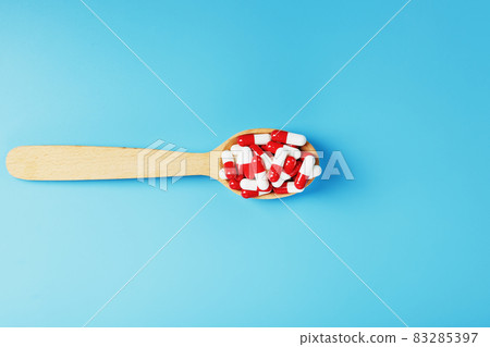 A handful of Red and white pill capsules in a wooden spoon on a blue background 83285397