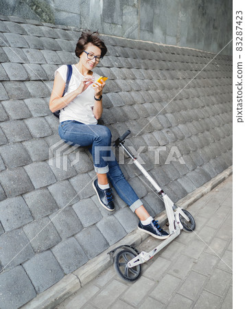 Smiling woman texting on smartphone after riding kick scooter. Female in jeans and sneakers on grey tiled wall background. Healthy lifestyle. Eco-friendly urban transport Smiling woman texting on smartphone after riding kick scooter. Female in jeans and sneakers on grey tiled wall background. Healthy lifestyle. Eco-friendly urban transport 83287423