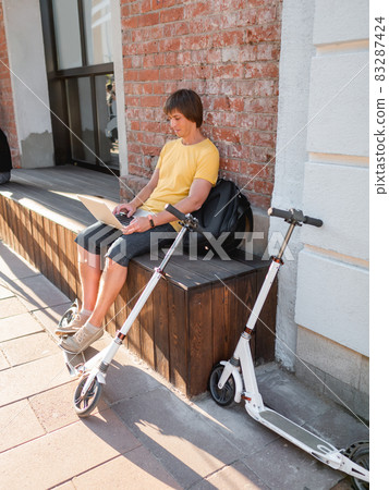 Man sits with laptop and kick scooter in street cafe. Freelancer at work. Student learns remotely from outdoors. Modern lifestyle. Summer vibes. Open air workplace. Man sits with laptop and kick scooter in street cafe. Freelancer at work. Student learns remotely from outdoors. Modern lifestyle. Summer vibes. Open air workplace. 83287424