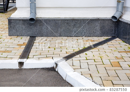 facade of a building wall with a downspout pipe and a concrete dish canal with iron grid on a stone slab sidewalk to drain rainwater near asphalt road city street, nobody. facade of a building wall with a downspout pipe and a concrete dish canal with iron grid on a stone slab sidewalk to drain rainwater near asphalt road city street, nobody. 83287550