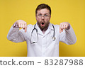 Agitated doctor in a white coat with a stethoscope around neck points down with index fingers, looks 83287988