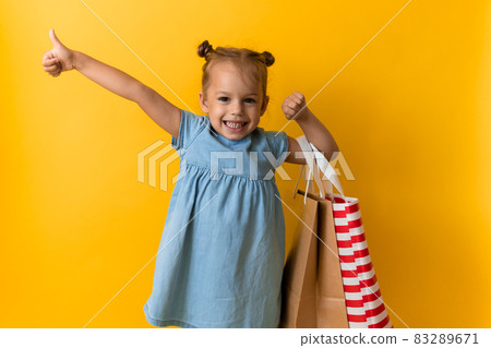 Portrait Caucasian Beautiful Happy Little Preschool Girl Smiling Cheerful And Holding Cardboard Bags Thumb Up Isolated On Orange Yellow Background. Happiness, Consumerism, Sale People shopping Concept 83289671
