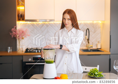 Portrait of attractive redhead young woman making healthy vegetable detox smoothie juice in blender standing in kitchen room, Portrait of attractive redhead young woman making healthy vegetable detox smoothie juice in blender standing in kitchen room, 83289856