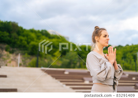 Side view of calm Caucasian young woman practicing yoga performing namaste pose outside in city park. 83291409