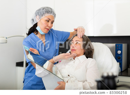 Young female cosmetologist attentive examines the face of an elderly female client before the procedure 83300258
