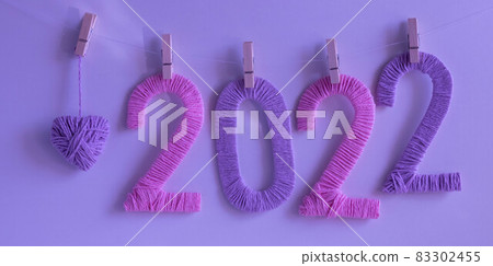 The numbers 2022, made of pink and lilac threads on a neon background, hang on clothespins next to a lilac knitted heart. The concept of the New Year The numbers 2022, made of pink and lilac threads on a neon background, hang on clothespins next to a lilac knitted heart. The concept of the New Year 83302455
