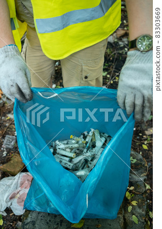 volunteer cleaning up used syringe in park and standing by bunch of garbage bag 83303669