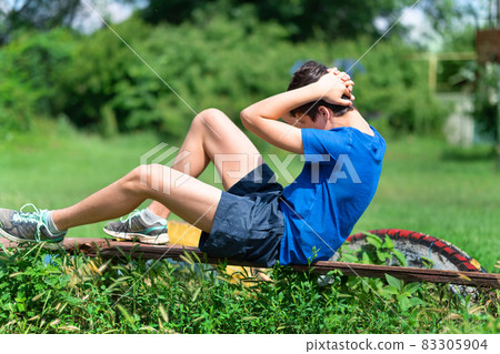 teenage boy exercising outdoors, sports ground in the yard, he pumps his abs on the board on the sports ground, healthy lifestyle 83305904