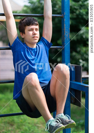 teenage boy exercising outdoors, sports ground in the yard, he hangs on the horizontal bar, raises his legs and pumps his abs, healthy lifestyle 83305905