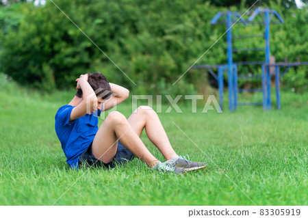 teenage boy exercising outdoors, sports ground in the yard, he pumps his abs on the sports ground green grass, healthy lifestyle teenage boy exercising outdoors, sports ground in the yard, he pumps his abs on the sports ground green grass, healthy lifestyle 83305919