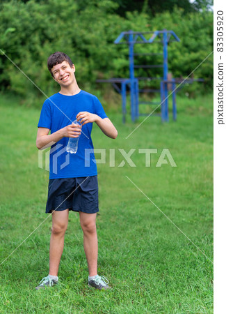 teenage boy exercising outdoors, sports ground in the yard, he opens a bottle of water and drinks, healthy lifestyle teenage boy exercising outdoors, sports ground in the yard, he opens a bottle of water and drinks, healthy lifestyle 83305920