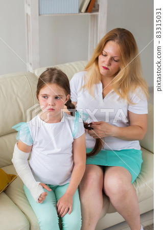 Mother braids a braid your child girl with a cast on a broken wrist or arm smiling. Childhood illnesses, a positive outlook and recovery. 83315031
