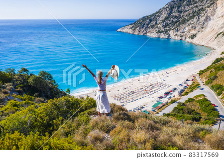 Happy woman standing on top of a rock, raising hands with an exciting feeling of freedom, looking at Myrtos Beach. Cephalonia island, Greece Happy woman standing on top of a rock, raising hands with an exciting feeling of freedom, looking at Myrtos Beach. Cephalonia island, Greece 83317569