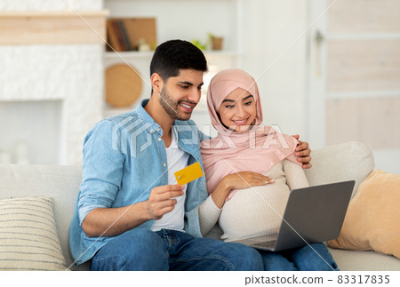 Preparing for childbirth. Loving muslim pregnant couple using laptop computer and credit card, sitting on sofa 83317835