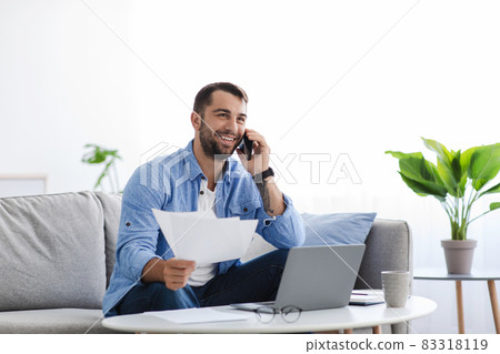 Busy smiling adult european guy sitting on sofa work at home with documents, talking with client by phone 83318119