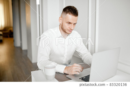 The manager works at home in the kitchen behind a laptop in a white shirt with a cup 83320136