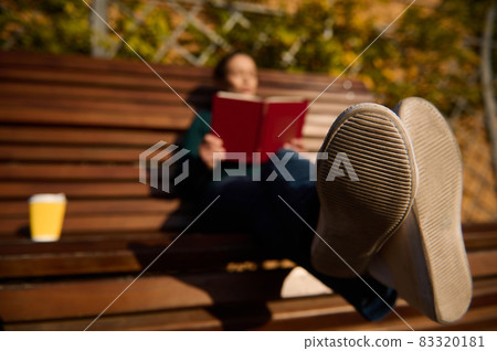 Main focus on the sole of sports sneakers of a blurred woman resting on a wooden bench in the park and reading a book and drinking coffee. Enjoying a weekend outdoor in autumn nature background 83320181