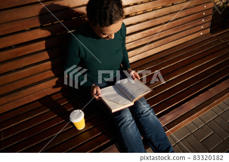 Overhead view of beautiful young brunette sitting on wooden bench in park, reading book and drinking coffee. Serene confident woman in casual denim with a book and takeaway paper cup of hot drink 83320182