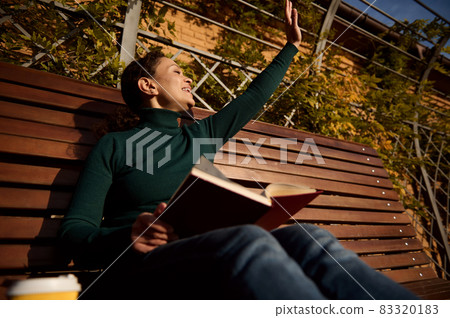 Relaxed contented young woman in casual denim greeting with hand, reading a book sitting on a wooden bench enjoying a warm sunny autumn weekend in country park, away from urban hustle 83320183