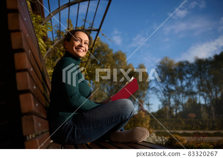 Gorgeous self-confident Hispanic woman in casual denim rests in a park sitting in lotus position on a wooden bench with book in her hands, smiles looking the side, enjoying weekend on autumn warm day 83320267
