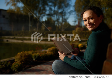 Attractive self-confident woman smiles looking through shoulder, reading book sitting on wooden bench on the background of beautiful lake in autumn nature background, resting away from urban bustle 83320268