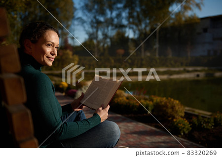 Portrait of a contented pretty woman sitting on wooden bench in park, reading book, enjoying warm sunny autumn day , away from the hustle and bustle of city, resting from digital gadgets and work 83320269
