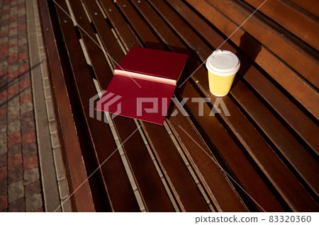High angle view of an open book with hard cover and a yellow takeaway cardboard paper cup with hot drink on a wooden bench in the park. Concept of outdoor rest away from gadgets and city bustle 83320360