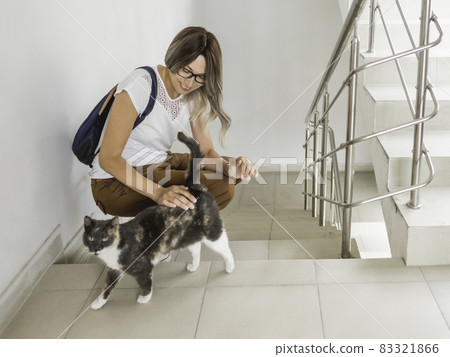 Smiling woman strokes stray cat on stairs. White staircase in apartment building. Smiling woman strokes stray cat on stairs. White staircase in apartment building. 83321866
