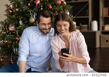 Happy young family couple using cellphone, sitting near Christmas tree. 83322508