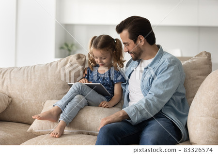 Caring smiling young father watching happy little daughter using tablet. 83326574