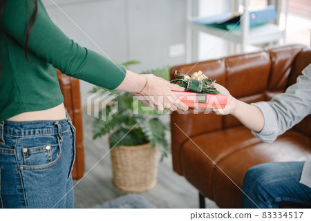 Close-Up of Couple Love Giving Present Together, Young Girl Surprise Gift to Her Boyfriend in The Living Room. Young Couple Love Give Present Box for Anniversary New Year Celebrate Together. 83334517