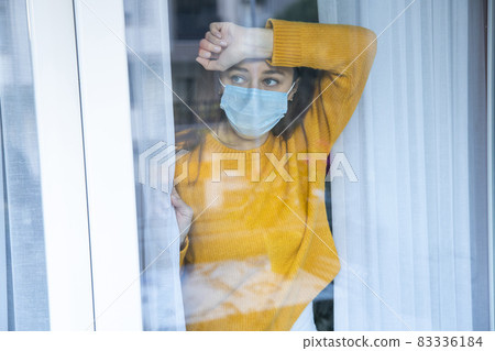 Young woman in quarantine wearing a mask and looking through the window 83336184