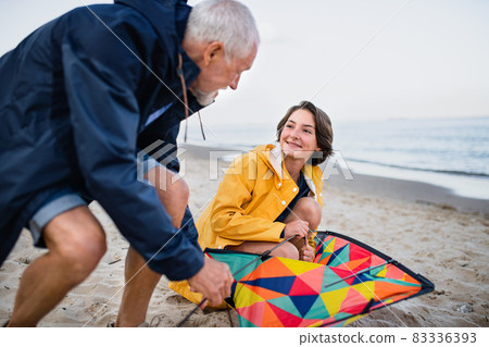 Senior man and his preteen granddaughter preparing kite for flying on sandy beach. Senior man and his preteen granddaughter preparing kite for flying on sandy beach. 83336393