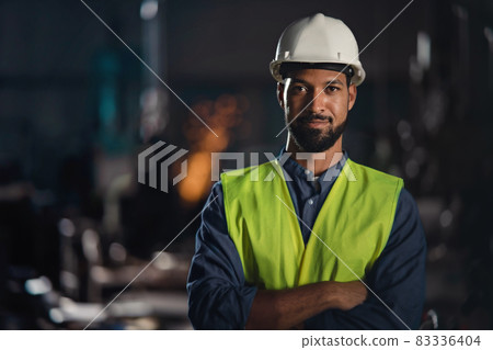 Portrait of happy young industrial man with protective wear indoors in metal workshop, looking at camera. Portrait of happy young industrial man with protective wear indoors in metal workshop, looking at camera. 83336404