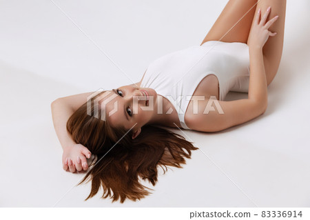 Cropped portrait of beautiful young woman in white bodysuit lying, posing isolated over gray background Cropped portrait of beautiful young woman in white bodysuit lying, posing isolated over gray background 83336914