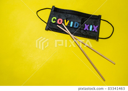 Black medical mask on a yellow background and Asian chopsticks in the shape of the letter V. The inscription COVID-19.  83341463