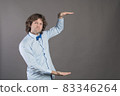 Horizontal shot of handsome Caucasian man with bristle, shows height of something with both hands, pretends holding item or thing, demonstrates size, wearing shirt and bow-tie, isolated on grey 83346264