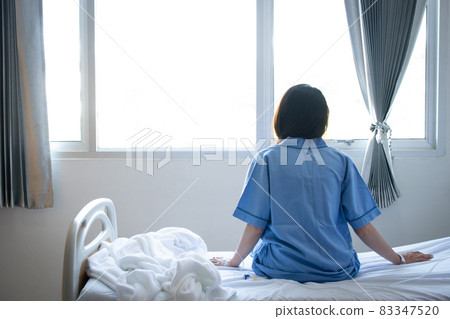 Back view of patient woman sitting on bed in hospital ward, looking away at window and hope everything will be better. Conceptual of patien lifestyle in hospital. 83347520