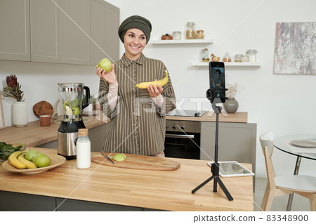 Young smiling woman showing fresh banana and apple to her online audience 83348900
