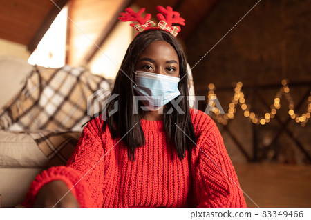 Serious young black lady wearing raindeer horns and face mask, staying home alone on Xmas during covid lockdown 83349466